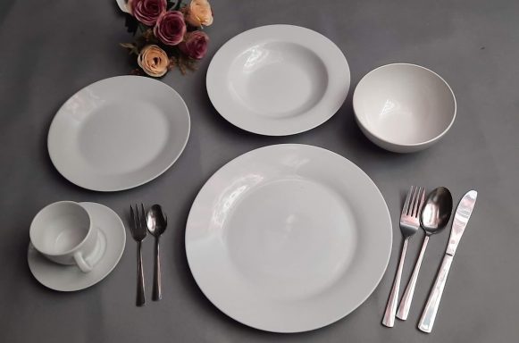 Tischgedeck mit weißen Tellern, Schalen, Besteck und einer Tasse auf grauem Hintergrund.